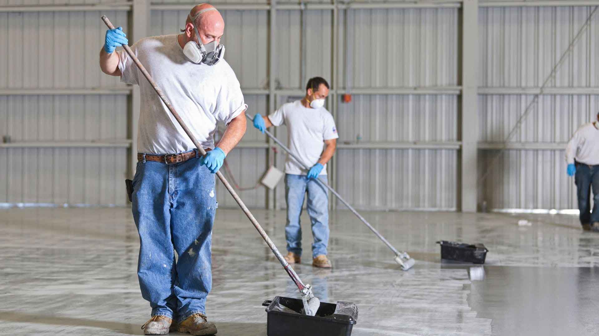 Workers in respirators applying grey epoxy coating across a large warehouse floor