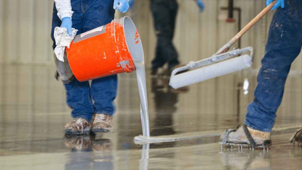 Tradesman pouring epoxy resin onto a warehouse floor while a colleague spreads it with a squeegee