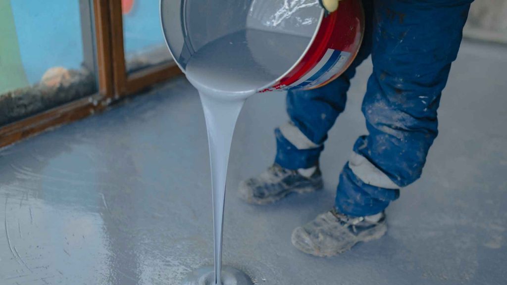 Tradesperson pouring liquid grey epoxy coating from a tin onto a floor