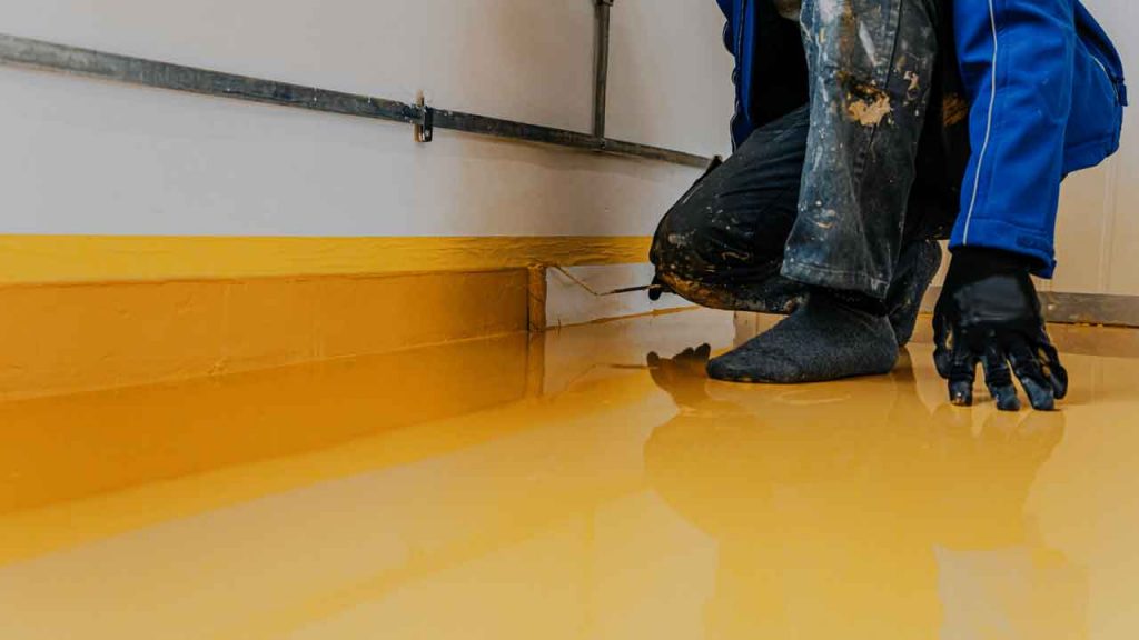 Worker applying a yellow self-levelling epoxy coating along a wall edge with a trowel